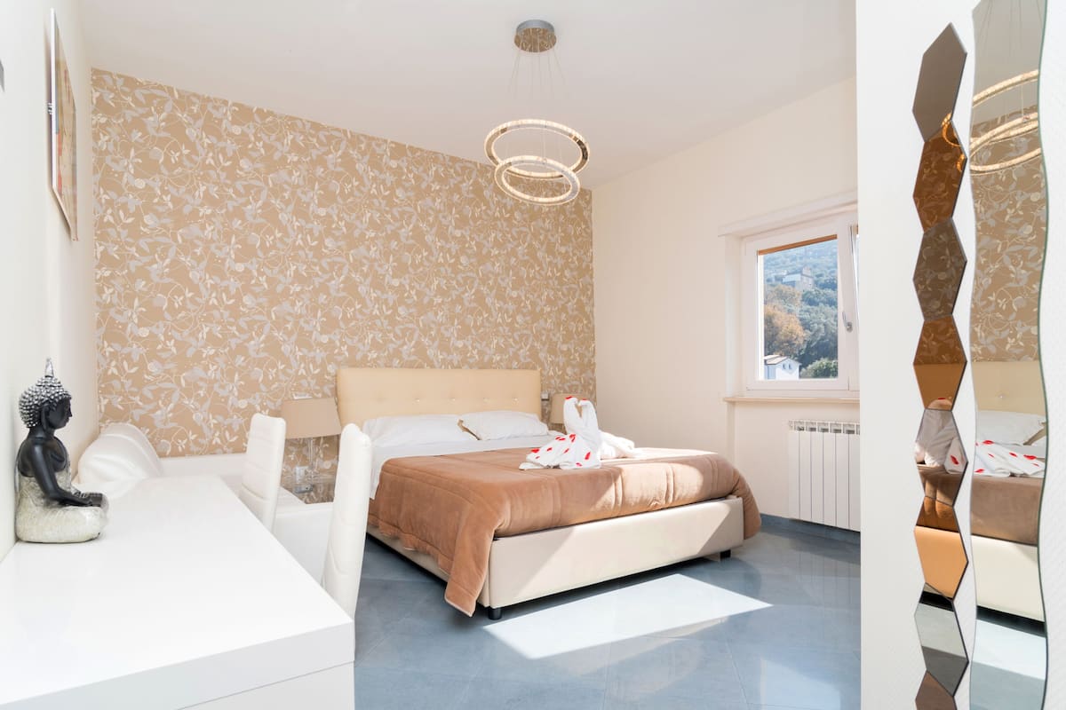 A modern bedroom with a large bed, a bedside table, a wall-mounted light fixture, and a window with a view.