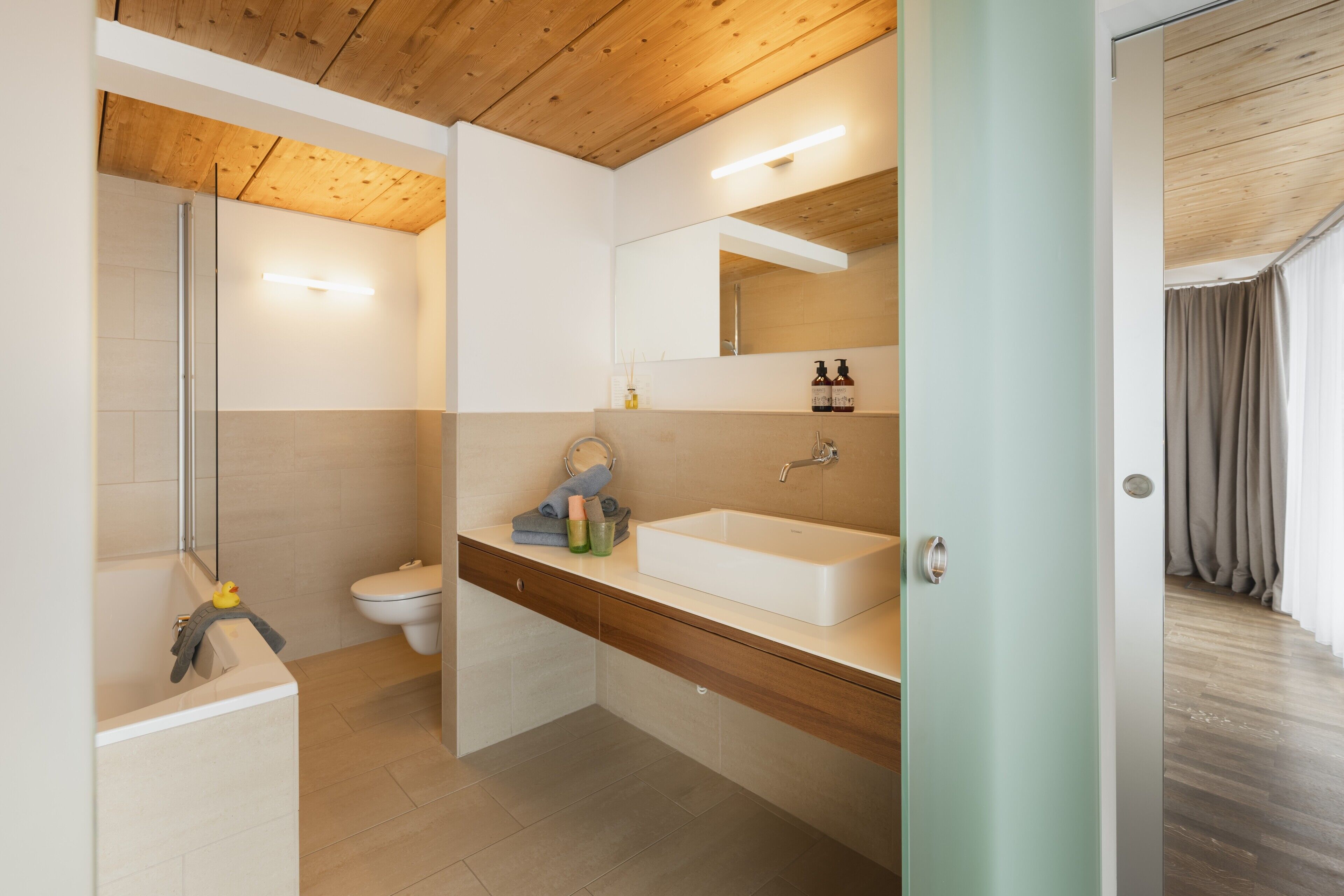 Een moderne badkamer met een houten plafond, een grote spiegel, een witte wastafel en een douchehoek.