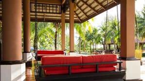 A covered outdoor seating area with red cushions, palm trees, and a pool in the background.