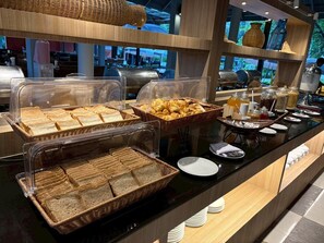 A buffet with various bread and pastries, clear plastic covers, and a counter with plates and condiments. - Palm Galleria Resort (Takua Pa)