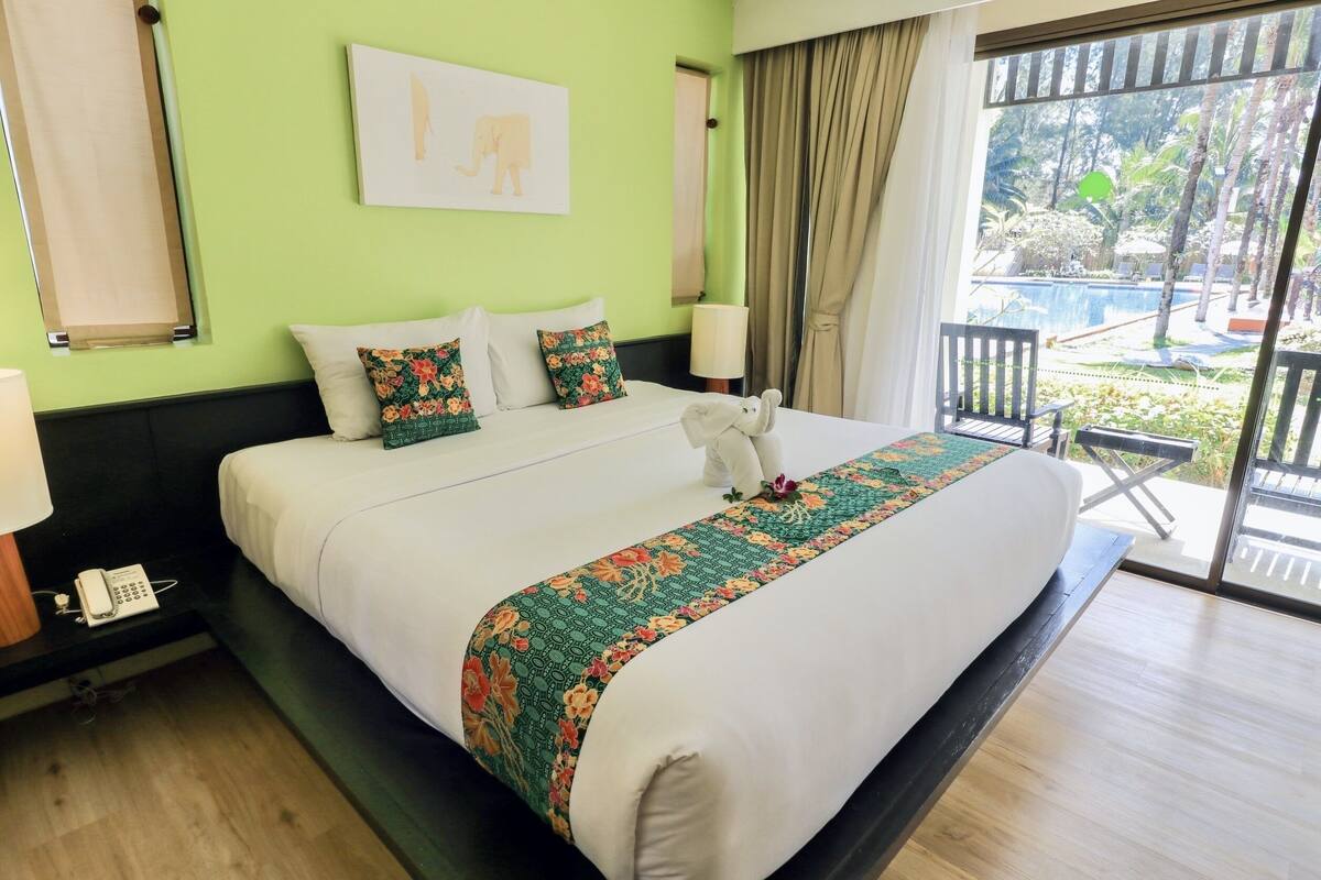 Hotelkamer met een groot bed, groene wanden, een raam met uitzicht op een zwembad en palmbomen, en een kleine tafel met een lamp.