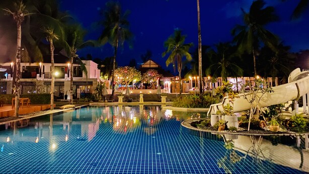 A nighttime view of a hotel pool area with a clear pool, palm trees, and a well-lit building in the background.