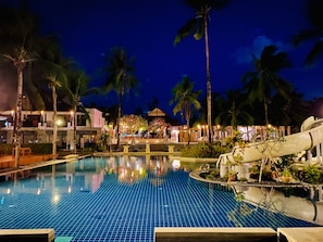 Vue nocturne d’une piscine d’hôtel, avec une eau cristalline, des palmiers et un bâtiment bien éclairé en arrière-plan.