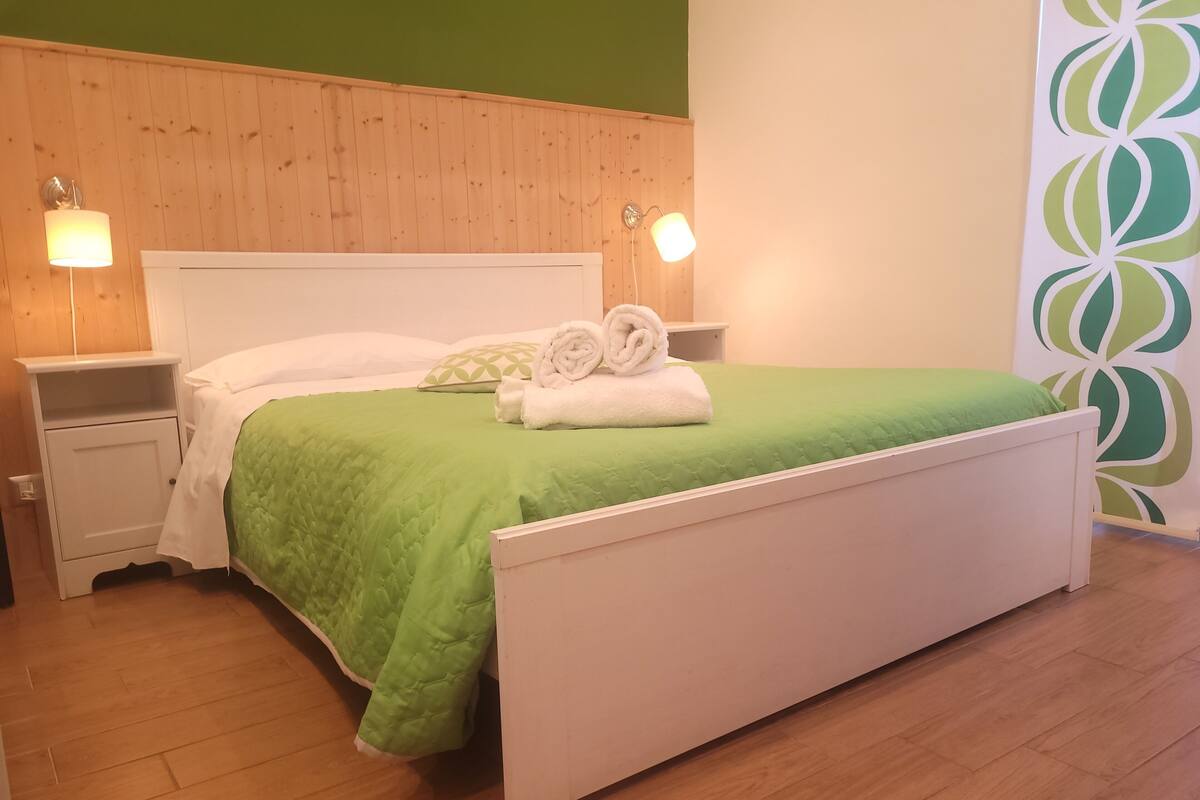 A neatly made bed with a green bedspread and two rolled towels, a white nightstand with a lamp, and a wooden headboard.