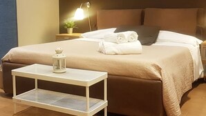 Cama bem arrumada com esquema de cores branco e castanho, uma pequena mesa branca com um candeeiro e um abajur numa mesa de cabeceira.