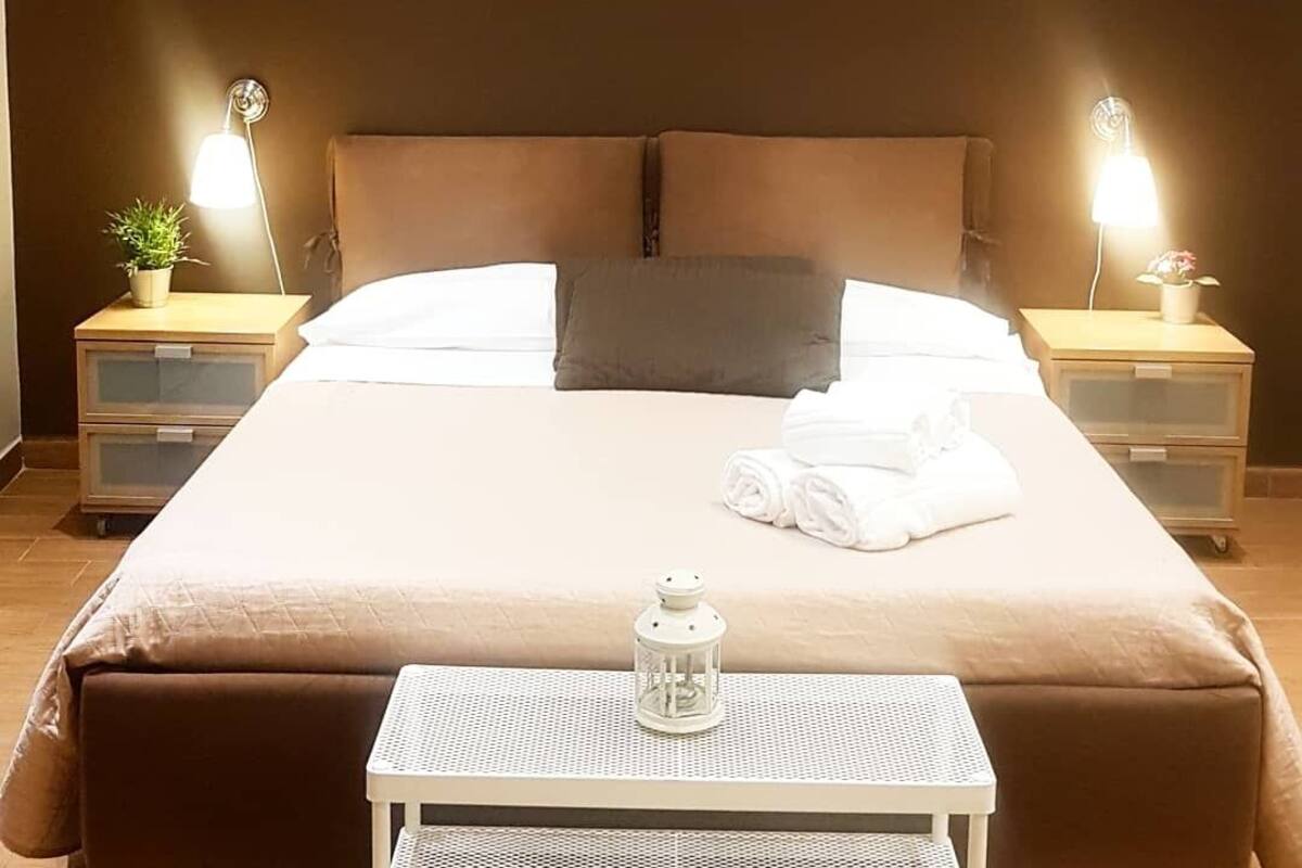 A neatly made bed with a pillow, two bedside tables with lamps, a small table with a lantern, and a potted plant on one of the tables.