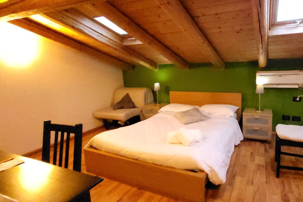 A room with a bed, bedside table, chair, and a small table with a lamp.