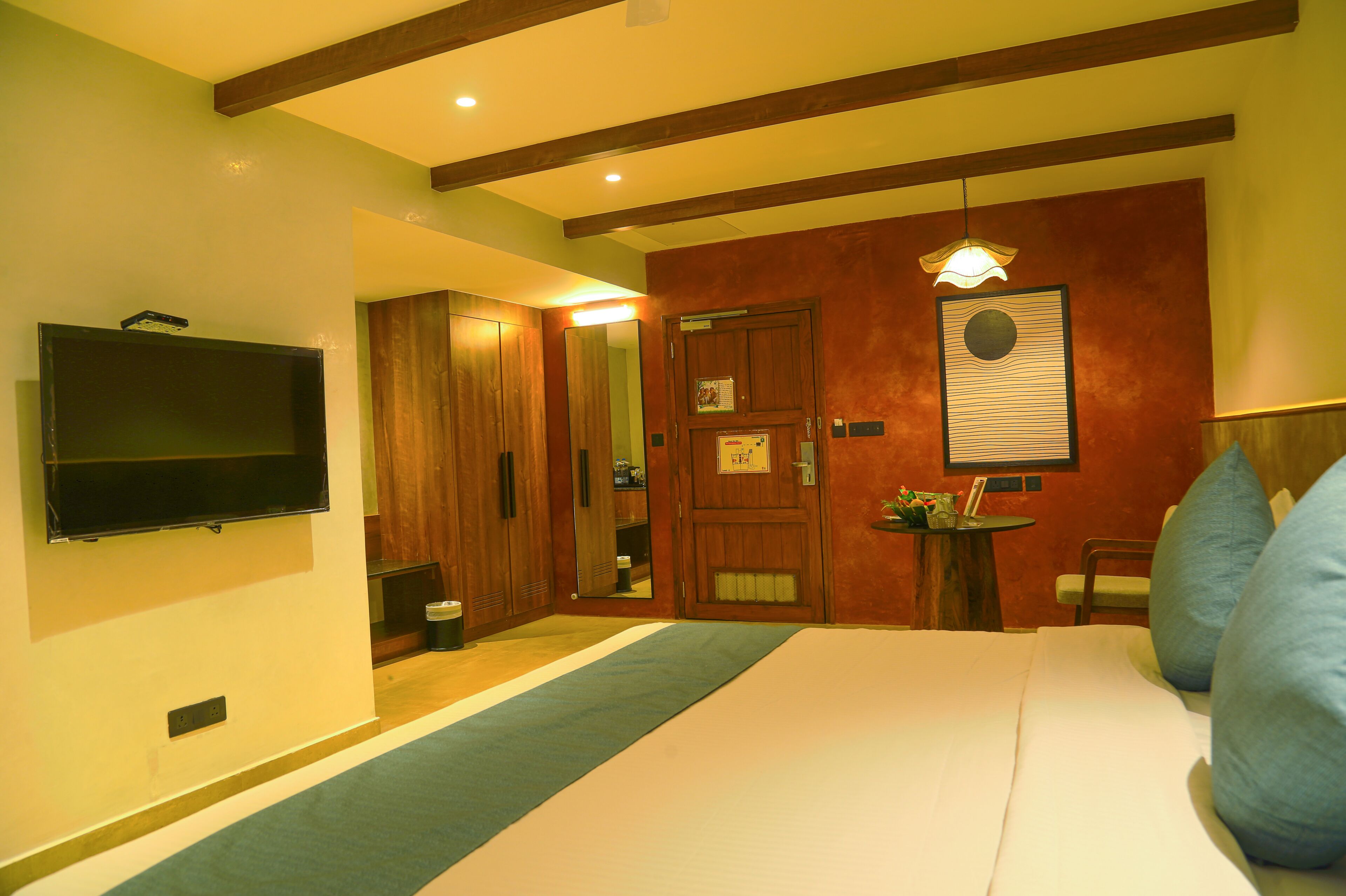 A hotel room with a bed, a television mounted on the wall, a wooden wardrobe, a small table with a lamp, and a mirror.