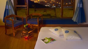Habitación de hotel con cama, dos almohadas, mesita de noche con un libro, una silla, una pequeña mesa con un farol, y un ventanal con vistas a la ciudad.