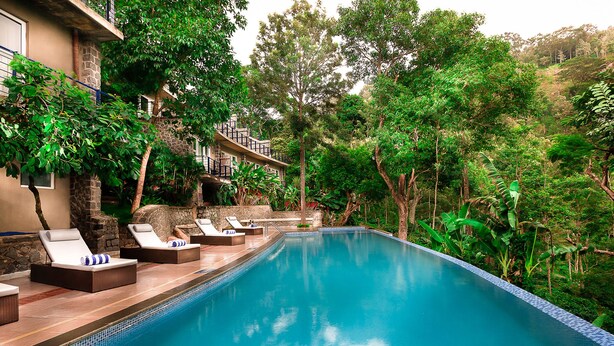 Piscina con sillones de descanso, rodeada de abundante vegetación y un edificio de piedra con balcón.