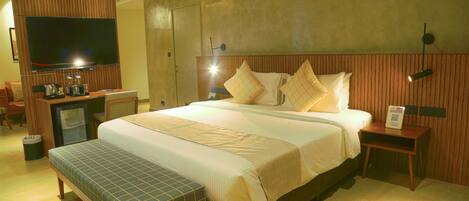 A hotel room with a large bed, a bench, a TV, a desk with a kettle, and a small refrigerator.