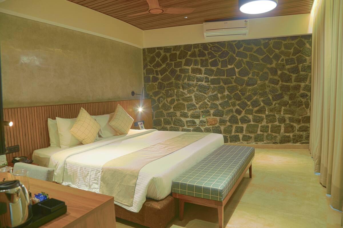 A hotel room with a stone wall, a wooden ceiling, a bed with a bench, and a small table with a kettle.