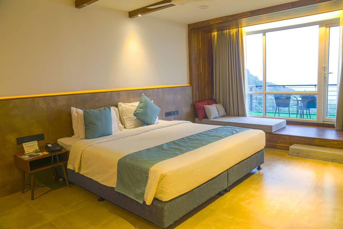 A hotel room with a large bed, bedside tables, a view of the outdoors, and a balcony with chairs.