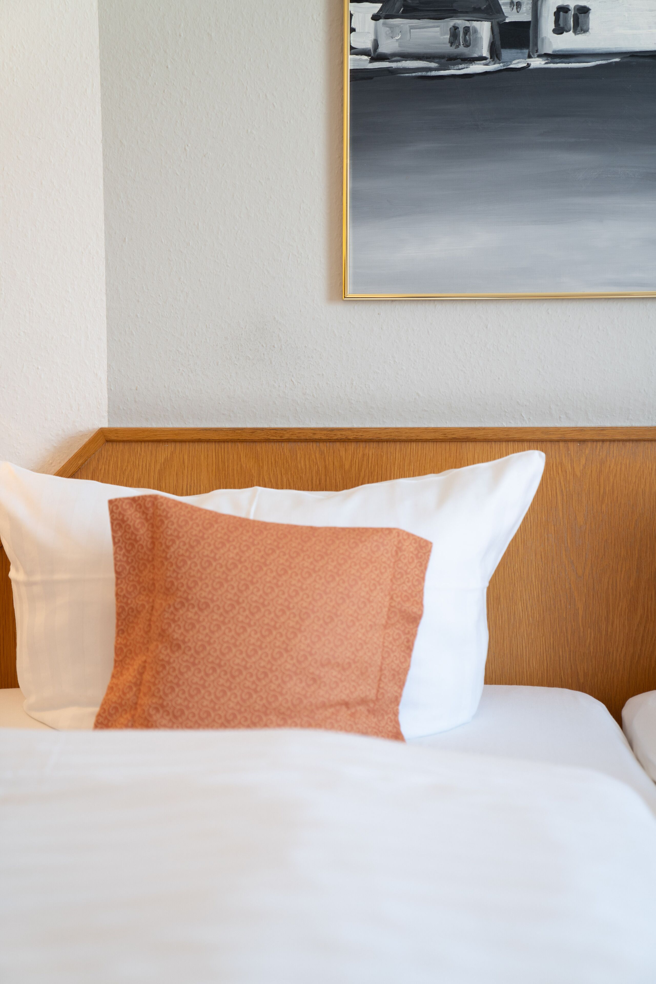 Habitación de hotel con cabecera de madera, cama con almohadas blancas y anaranjadas, y un cuadro blanco y negro en la pared.