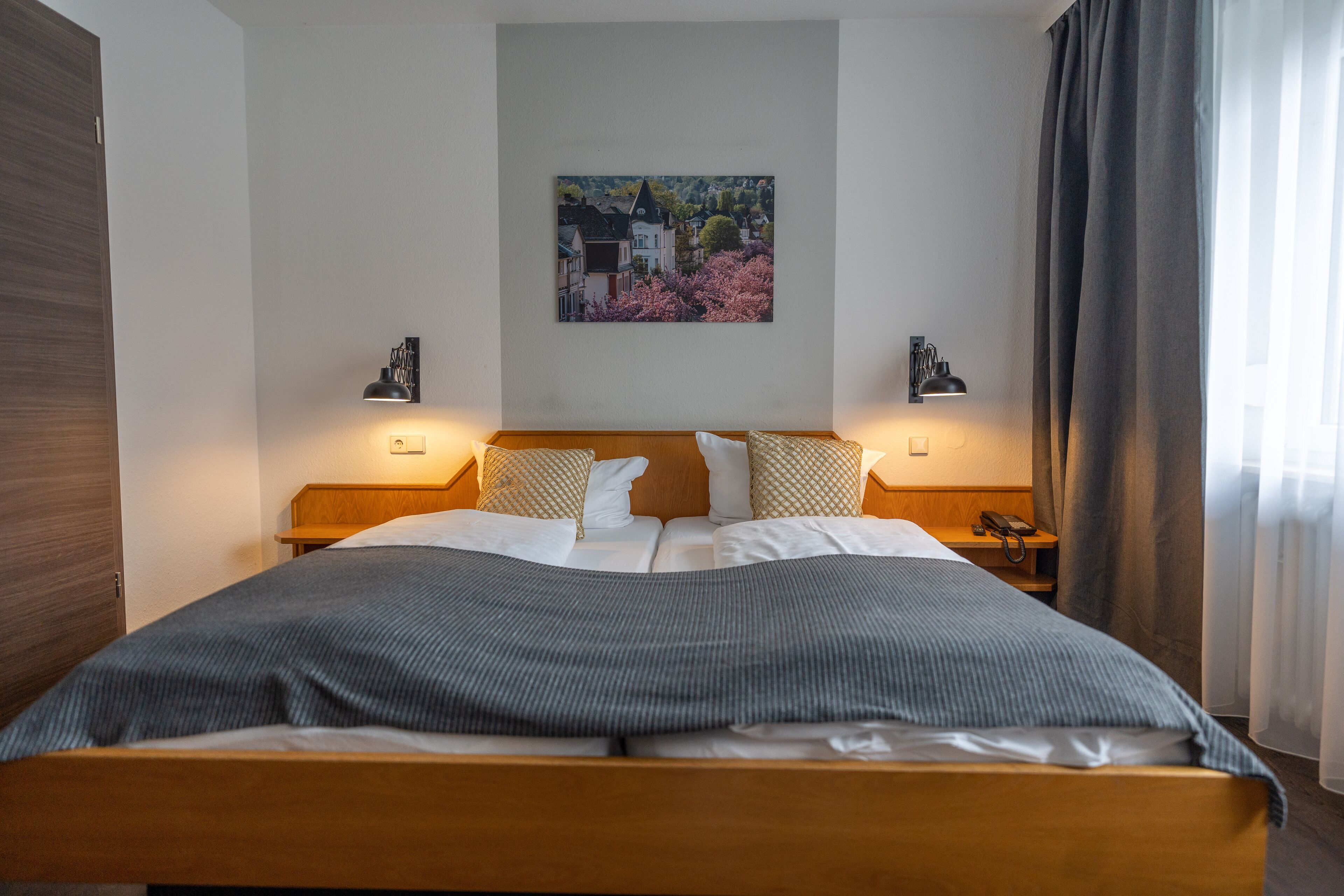 Un lit bien fait, recouvert d’une couverture grise, avec deux lampes de chevet, un tableau accroché au mur et une tête de lit en bois.