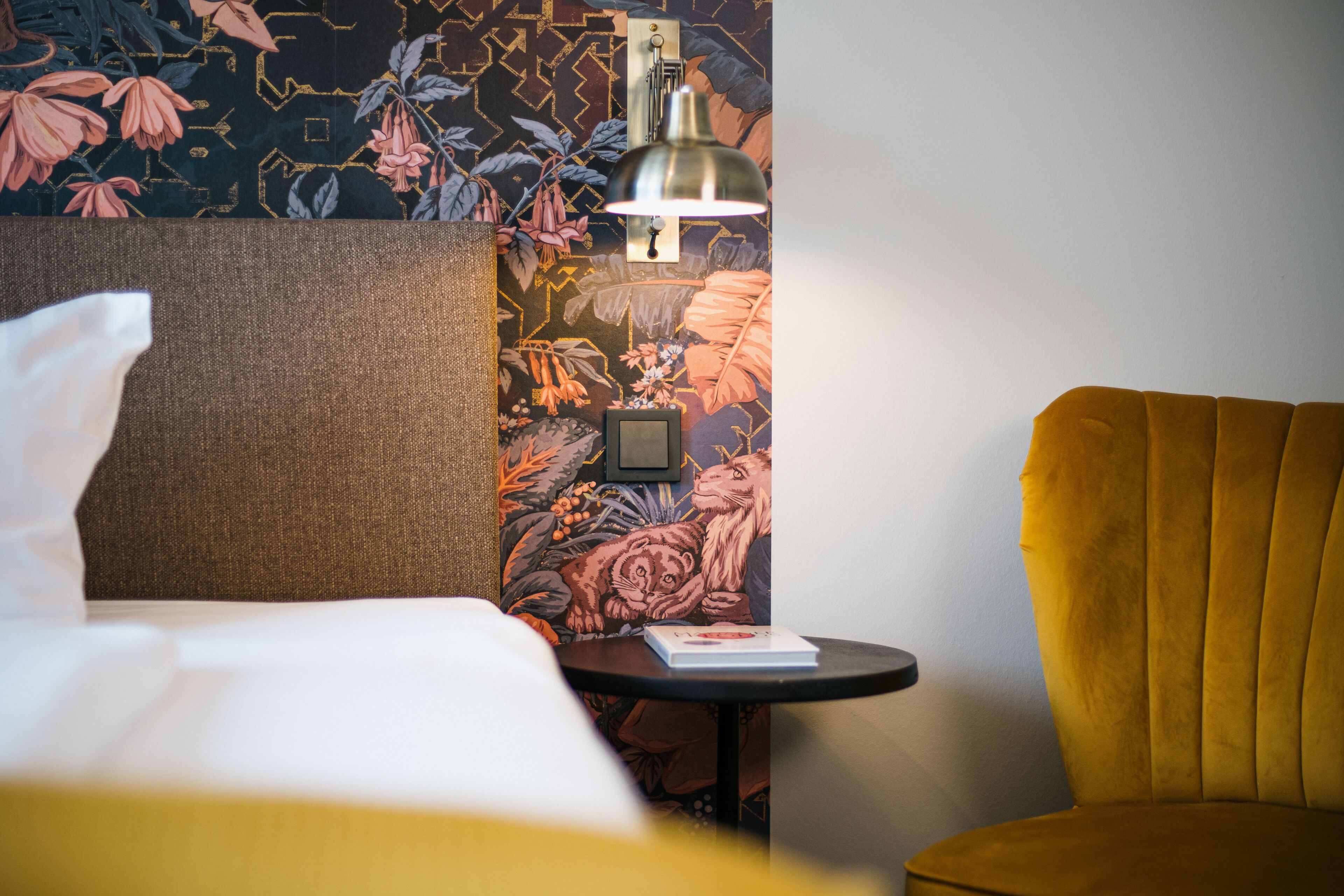 Una habitación de hotel con una cama, una mesita con un libro, una lámpara y un sillón amarillo.