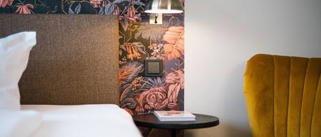 Una habitación de hotel con una cama, una mesita con un libro, una lámpara y un sillón amarillo.