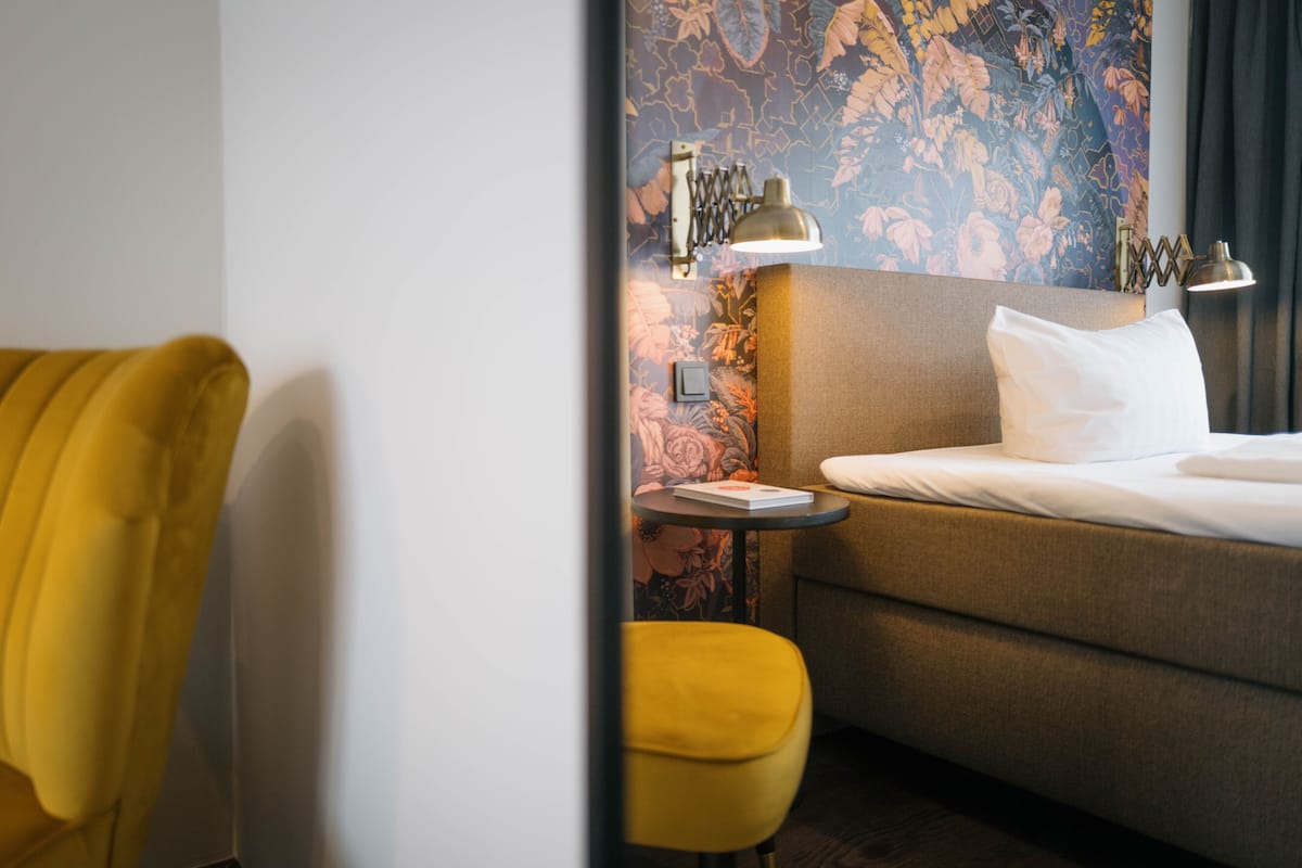 A hotel room with a bed, a yellow armchair, a small table with a lamp, and a floral wallpaper.
