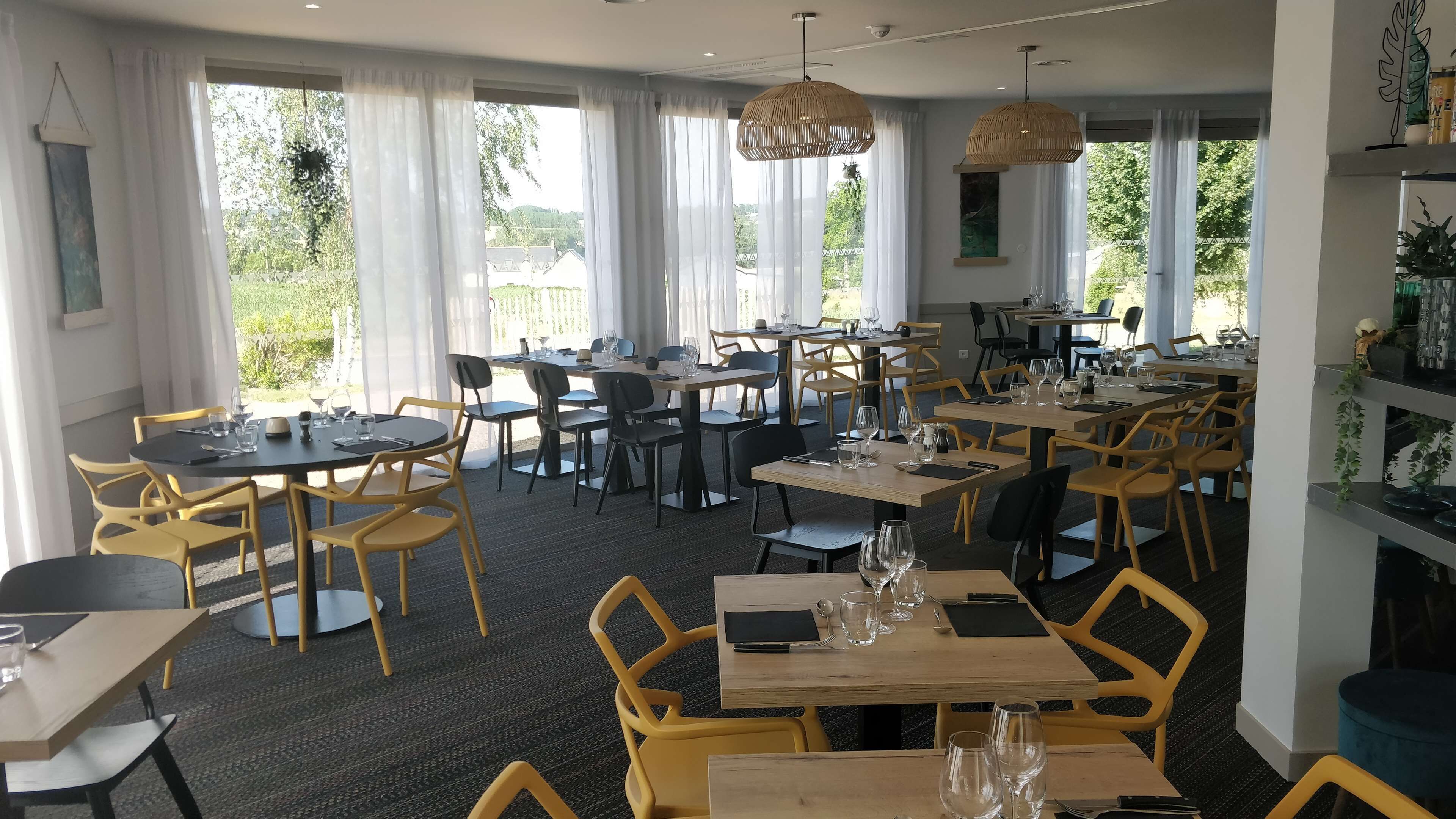 A restaurant with multiple tables and chairs, large windows with white curtains, and hanging rattan light fixtures.