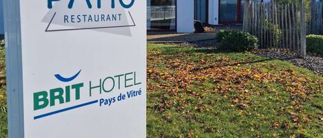 A sign for Le Patio Restaurant and Brit Hotel in a modern building with large windows and a wooden fence.