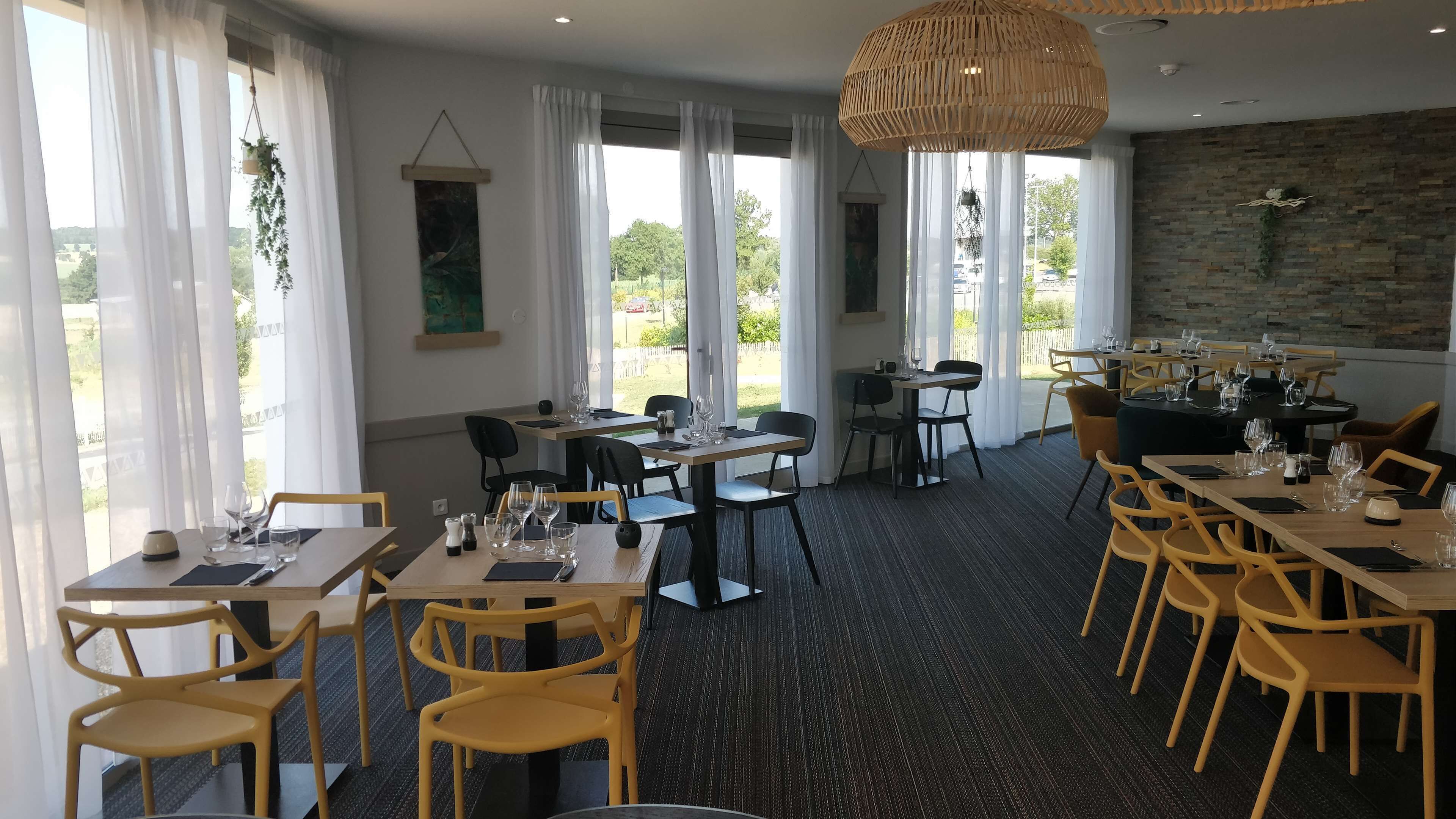 Un restaurante con varias mesas y sillas, ventanas amplias con cortinas blancas, una lámpara colgante y una pared de piedra.