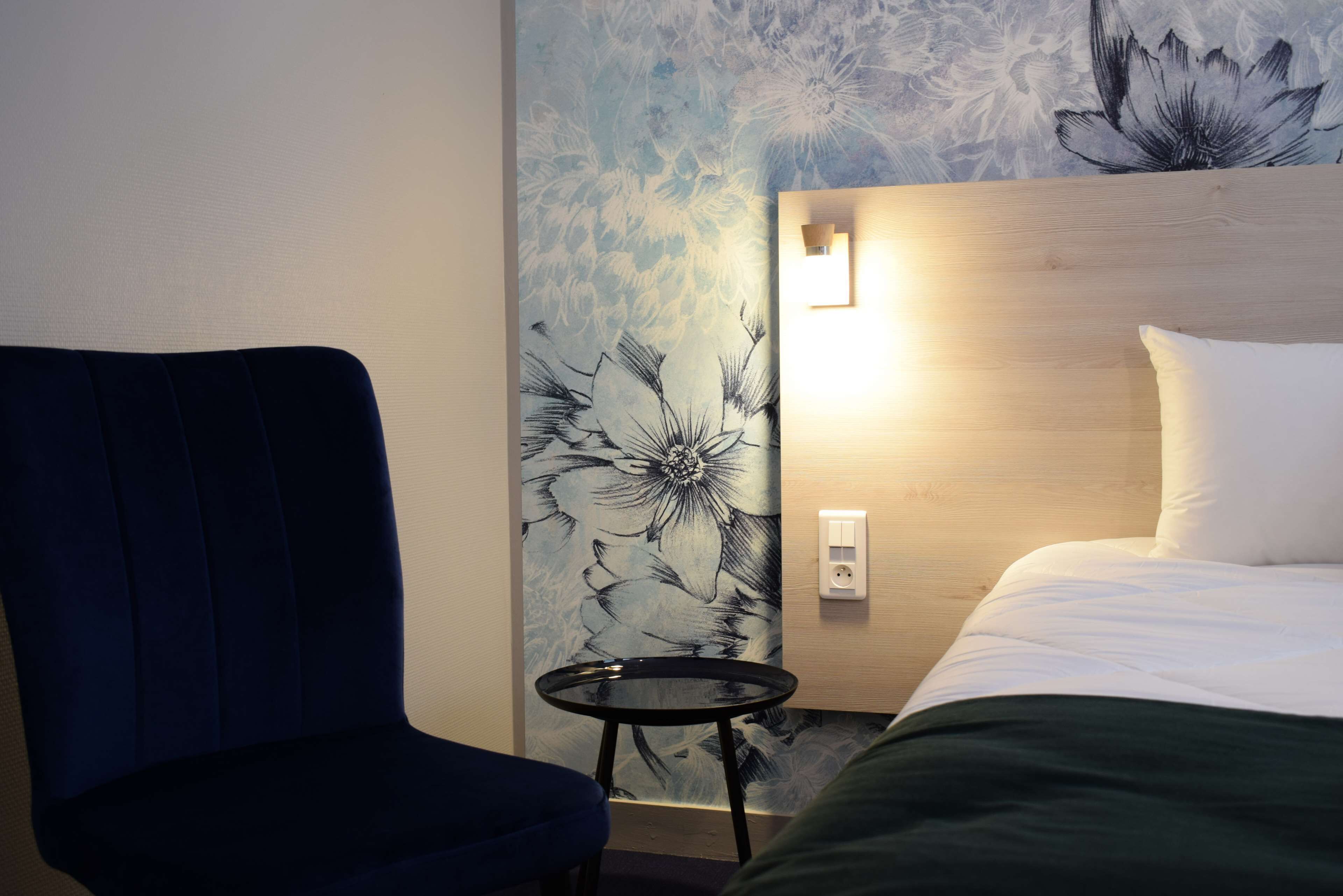 A hotel room with a bed, a blue armchair, a small round table, and a floral wallpaper.