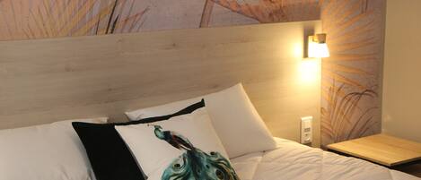 Una cama bien tendida con ropa blanca y almohadas decorativas, incluyendo una con diseño de pavo real, frente a una pared con un mural floral texturizado.