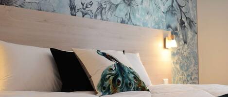 Ein Bett mit weißer Bettwäsche und einem türkisblauen Pfauen-Kissen, ein Holz-Kopfteil und eine Wand mit floralem Muster.