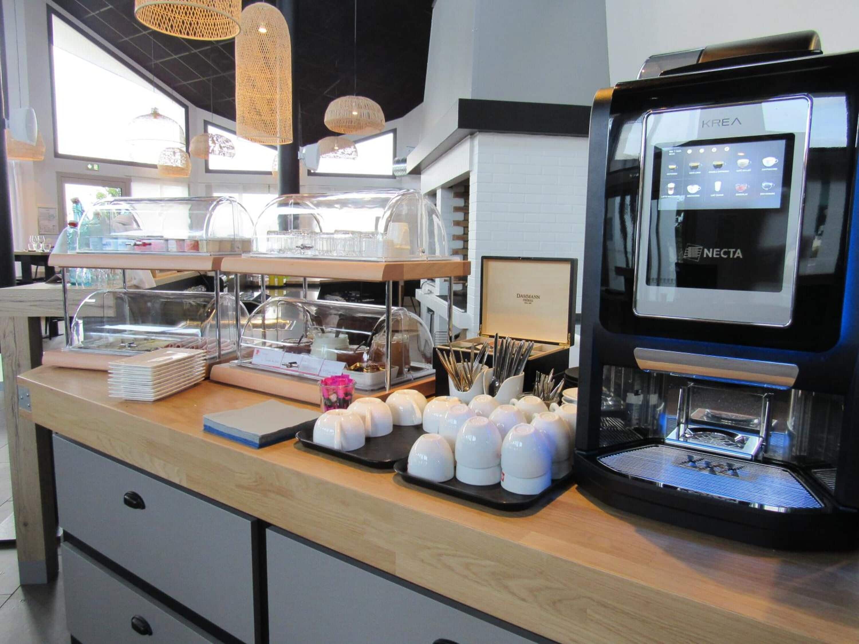 Un espace dédié au café, comprenant une machine à café Krea Necta, une vitrine réfrigérée et divers types de tasses et d’ustensiles à café.