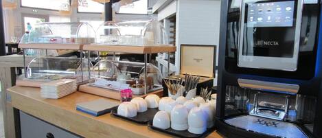 Un espace dédié au café, comprenant une machine à café Krea Necta, une vitrine réfrigérée et divers types de tasses et d’ustensiles à café.