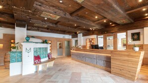 A reception area with a wooden reception desk, a white counter, and a bench. There is a fireplace with decorative items and a plant. The ceiling has exposed wooden beams and recessed lighting. - Hotel Untersberg (Groedig)