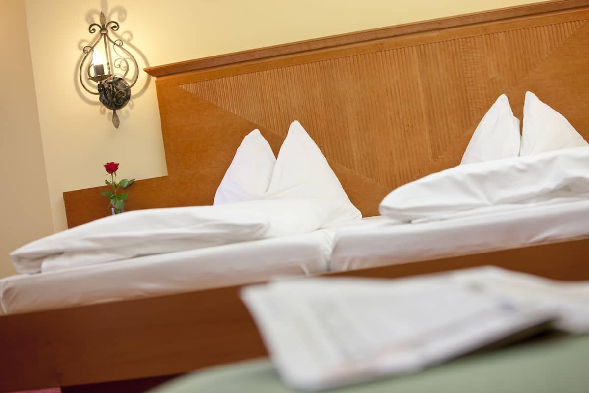 A neatly made bed with white linens and pillows, a wooden headboard, and a wall-mounted light fixture.