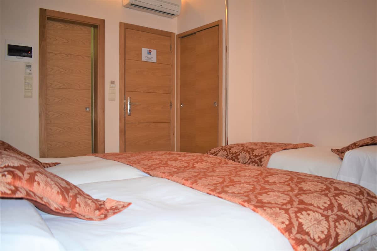 Habitación de hotel con dos camas, cada una con una colcha y almohadas estampadas. Hay puertas de madera con un cartel en una de ellas, y un equipo de aire acondicionado instalado en la pared.