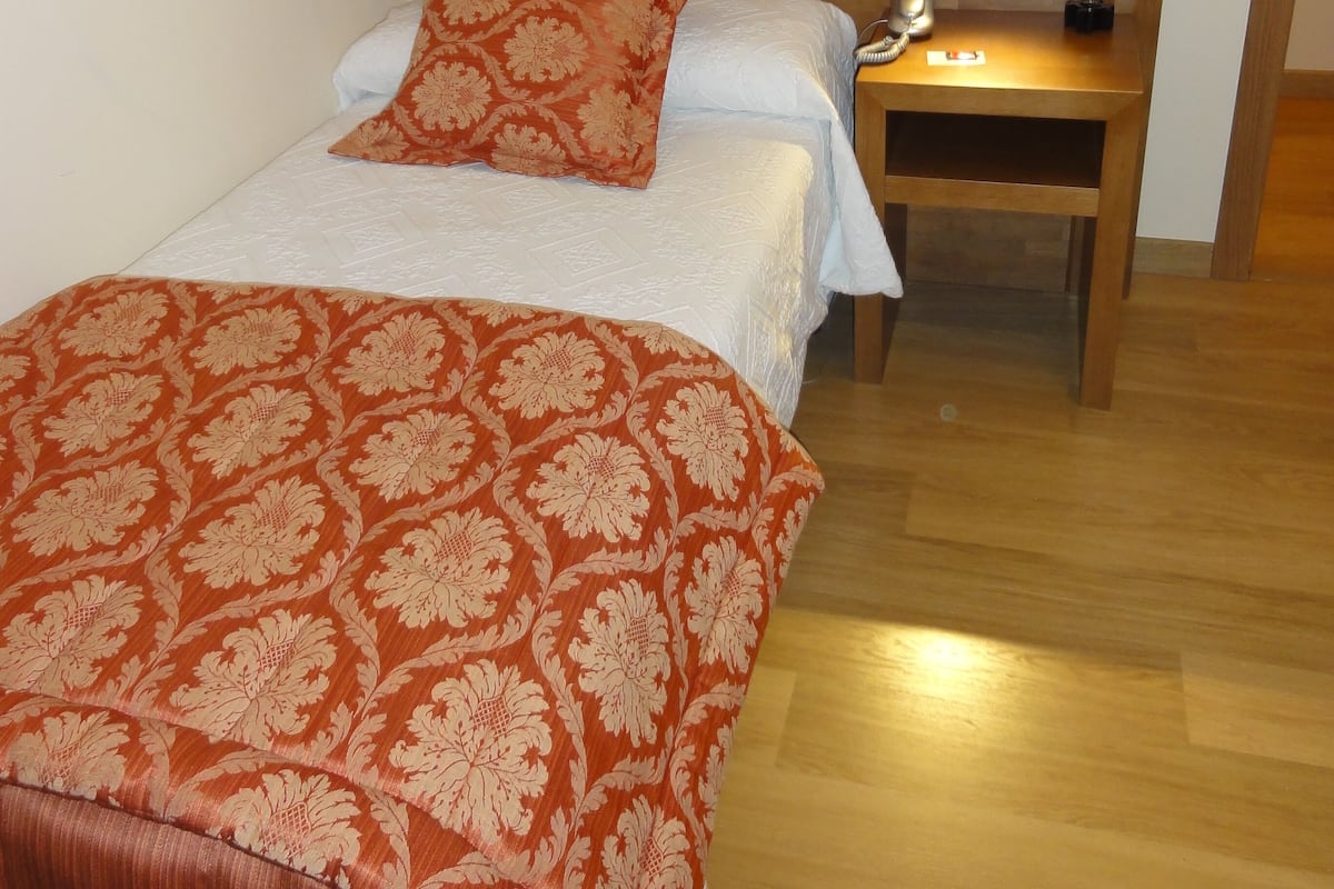 Un dormitorio con cabecera de madera, una cama con colcha floral roja, una mesita de noche con lámpara y una pequeña planta en maceta.