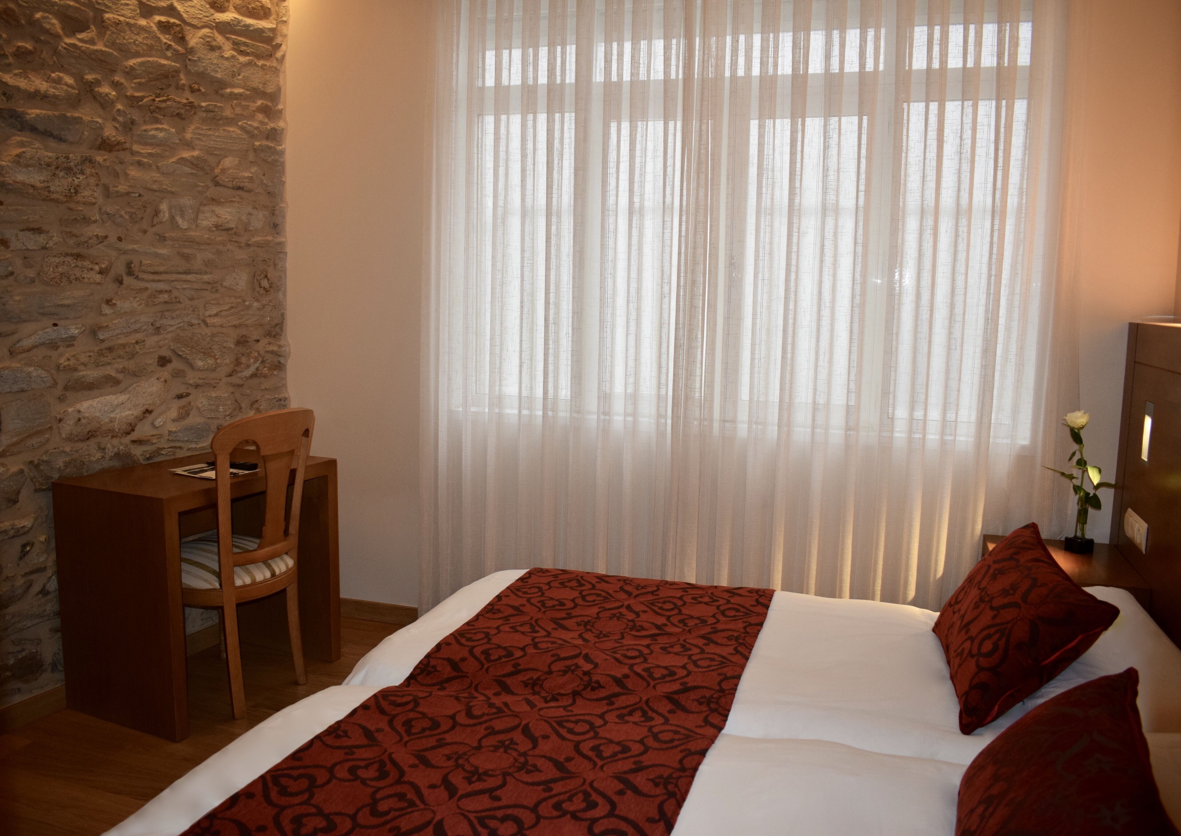 Un dormitorio con pared de piedra, una silla de madera, una cama con colcha estampada y una ventana con cortinas traslúcidas.