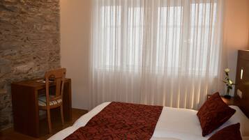 Un dormitorio con pared de piedra, una silla de madera, una cama con colcha estampada y una ventana con cortinas traslúcidas.