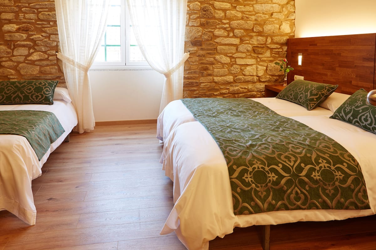 Habitación doble con dos camas, cada una con ropa de cama verde y blanca, pared de piedra, cabecero de madera y una ventana con cortinas traslúcidas.