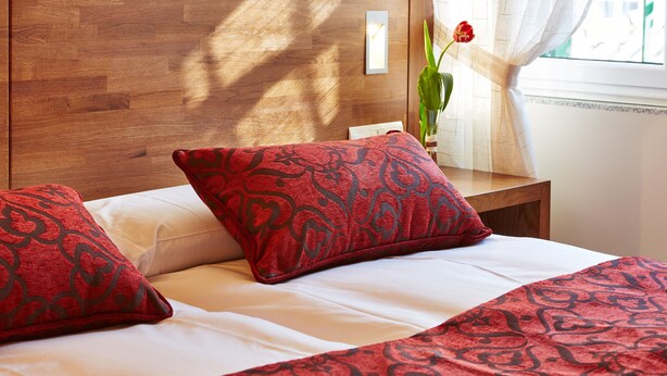 A neatly made bed with red and white bedding, a wooden headboard, and a vase with a red flower on a bedside table. - Mapoula (Santiago de Compostela)