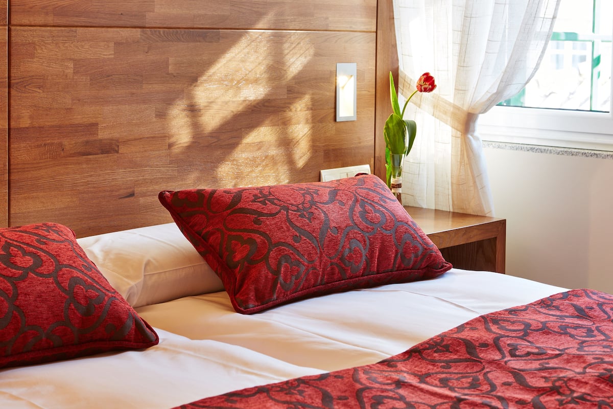 Una cama bien hecha con ropa de cama roja y blanca, un cabecero de madera y un jarrón con una flor roja sobre una mesita de noche.