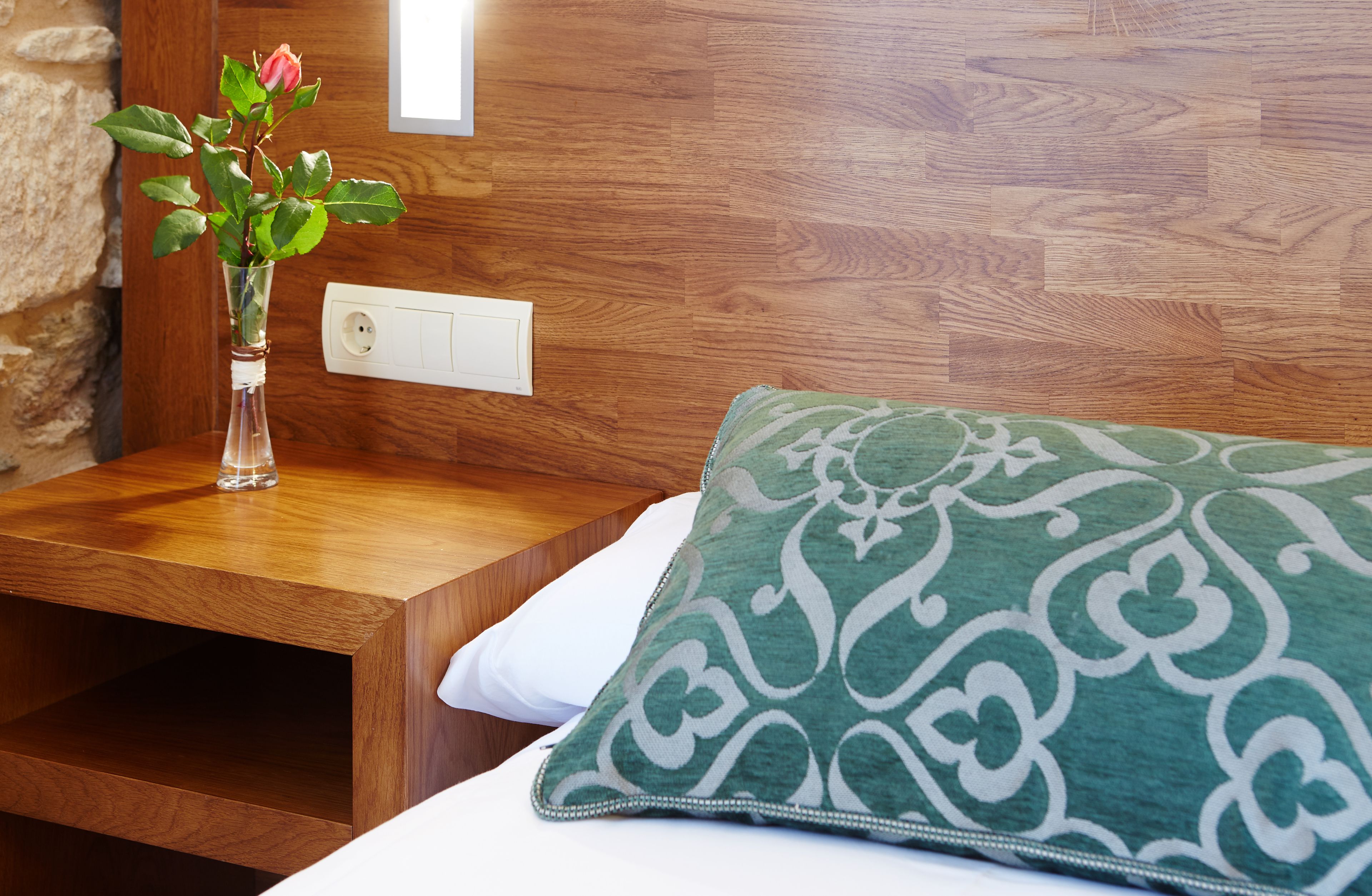 Una mesita de noche de madera con un florero y un cojín estampado verde sobre una cama.
