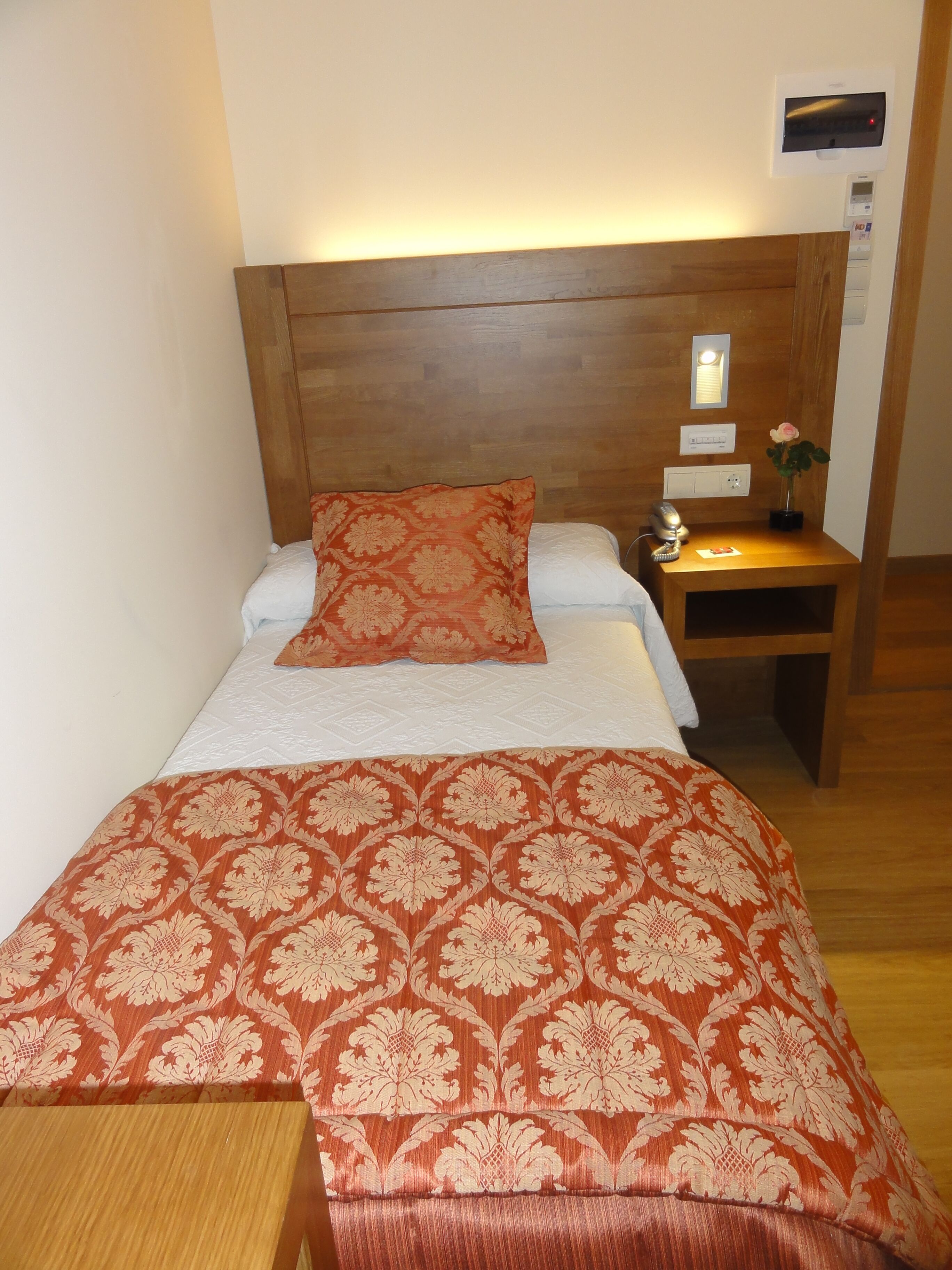 A neatly made bed with a patterned bedspread, a wooden headboard, a bedside table with a lamp, and a small vase with a flower.
