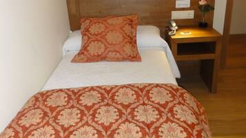 A neatly made bed with a patterned bedspread, a wooden headboard, a bedside table with a lamp, and a small vase with a flower.