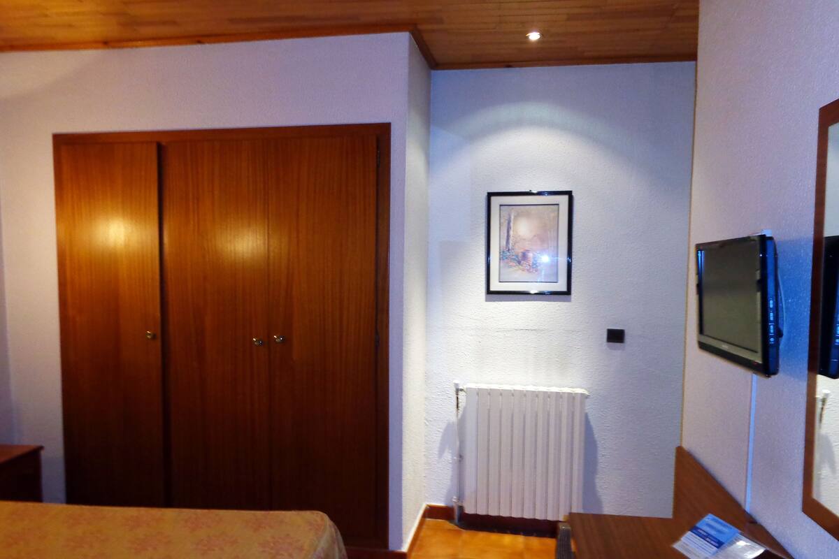 A hotel room with a wooden wardrobe, a framed picture on the wall, a television mounted on the wall, and a bedside table with a card.