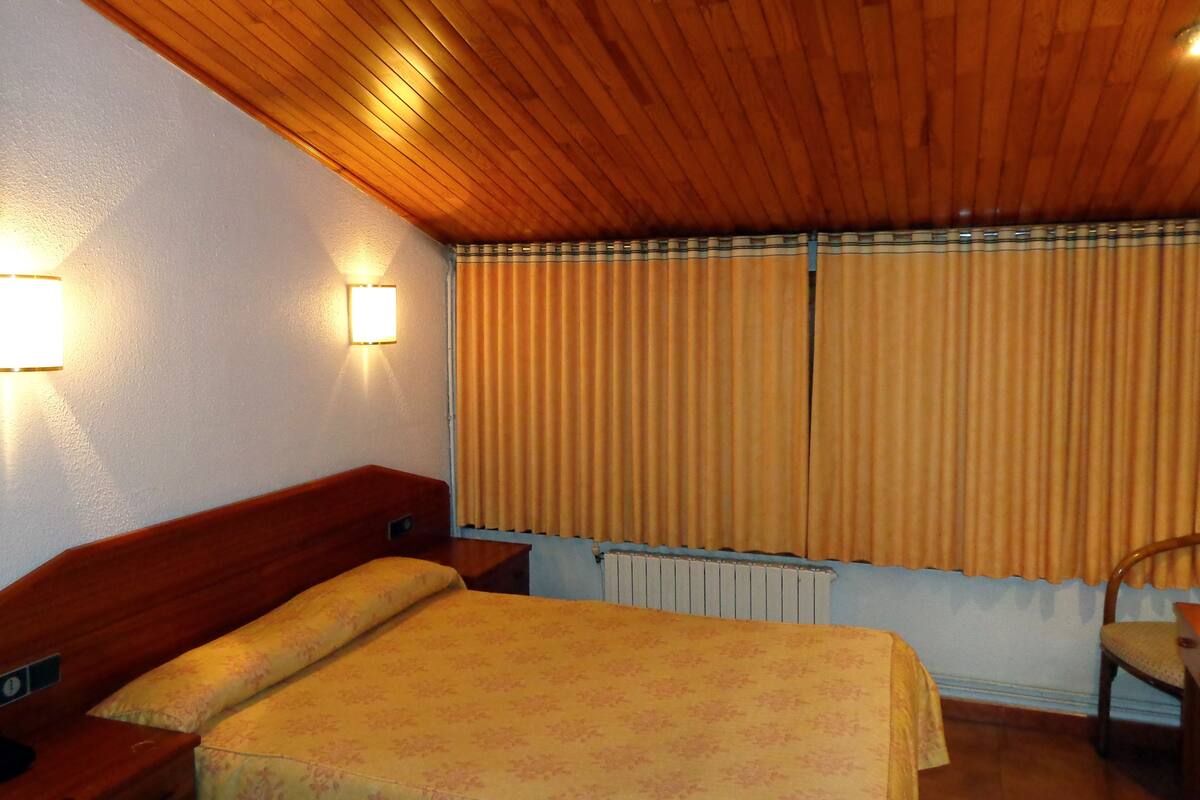 A room with a wooden ceiling, a bed with a floral bedspread, a nightstand, a chair, and a radiator.