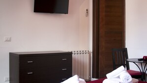 Ein Schlafzimmer mit Kommode, Wandfernseher, Heizkörper, Holztür und einem kleinen Tisch mit Stuhl.