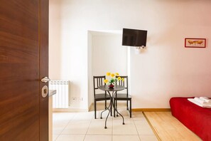 A hotel room with a wooden door, a small table with chairs, a red sofa, a flat-screen TV mounted on the wall, and a framed picture of birds. - Vatican City Guest House (Rome)