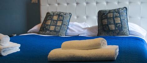 A neatly made bed with blue bedding and two decorative pillows, a white headboard, and a stack of white towels.