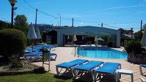 Un área de piscina con sillones reclinables azules y sombrillas, una zona de estar cubierta y un cielo azul despejado.