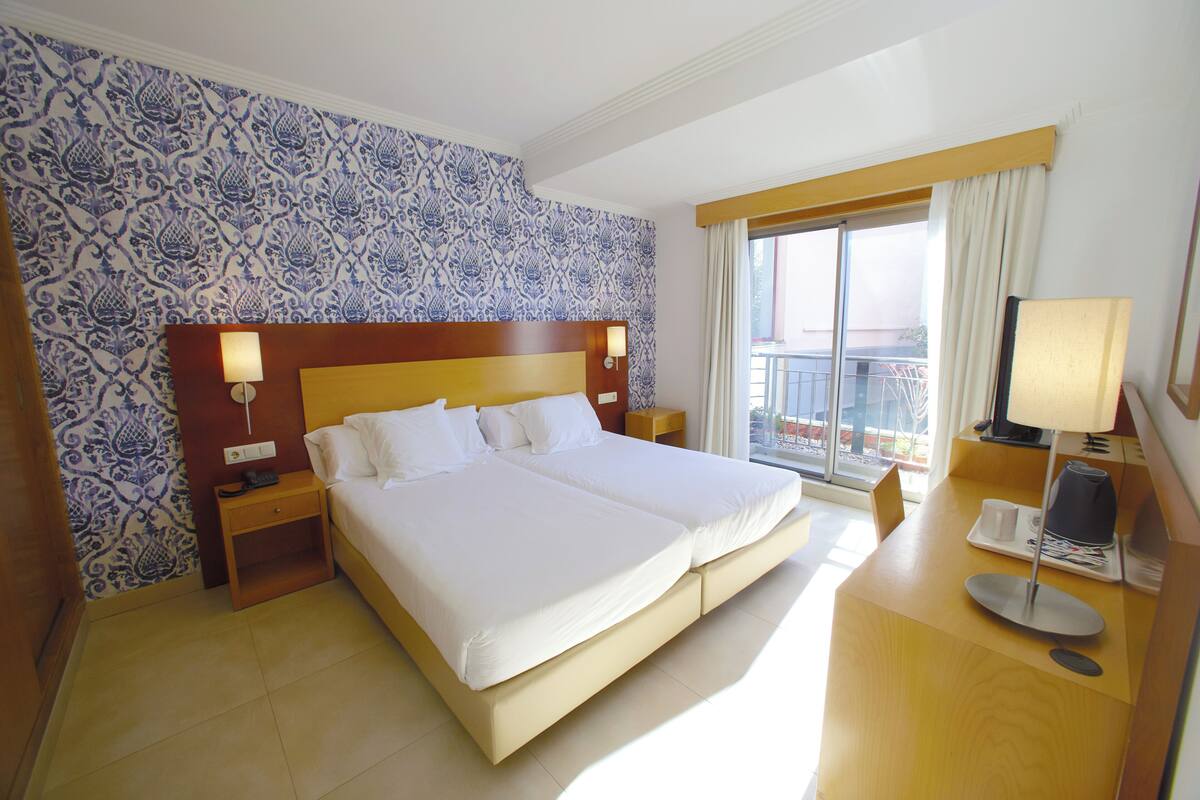 A hotel room with a large bed, two bedside tables with lamps, a desk with a computer, and a window with a view of the outside.
