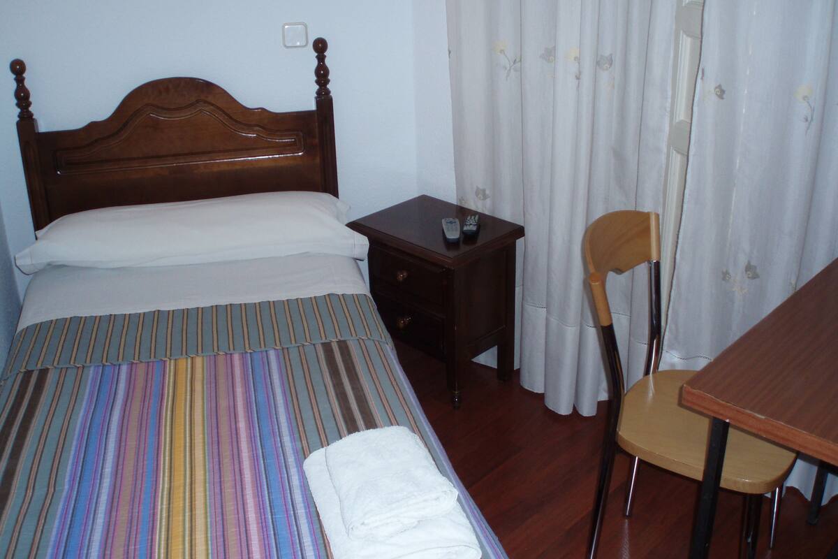 Ein Hotelzimmer mit einem hölzernen Bett, einer gestreiften Tagesdecke, einem weißen Kopfkissen, einem kleinen Nachttisch aus Holz mit einer Fernbedienung, einem Holzstuhl und einem weißen Vorhang.